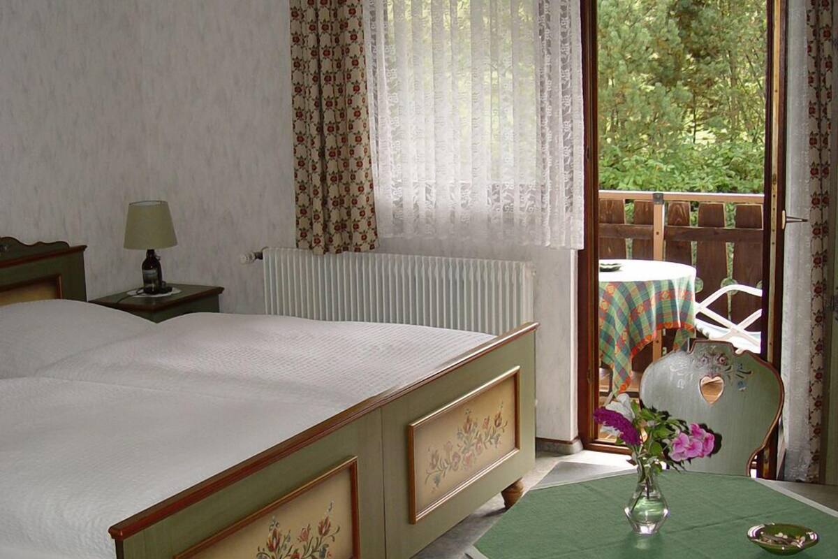 A bedroom with a bed, a table with a vase of flowers, a chair, a window with curtains, and a view of greenery outside.