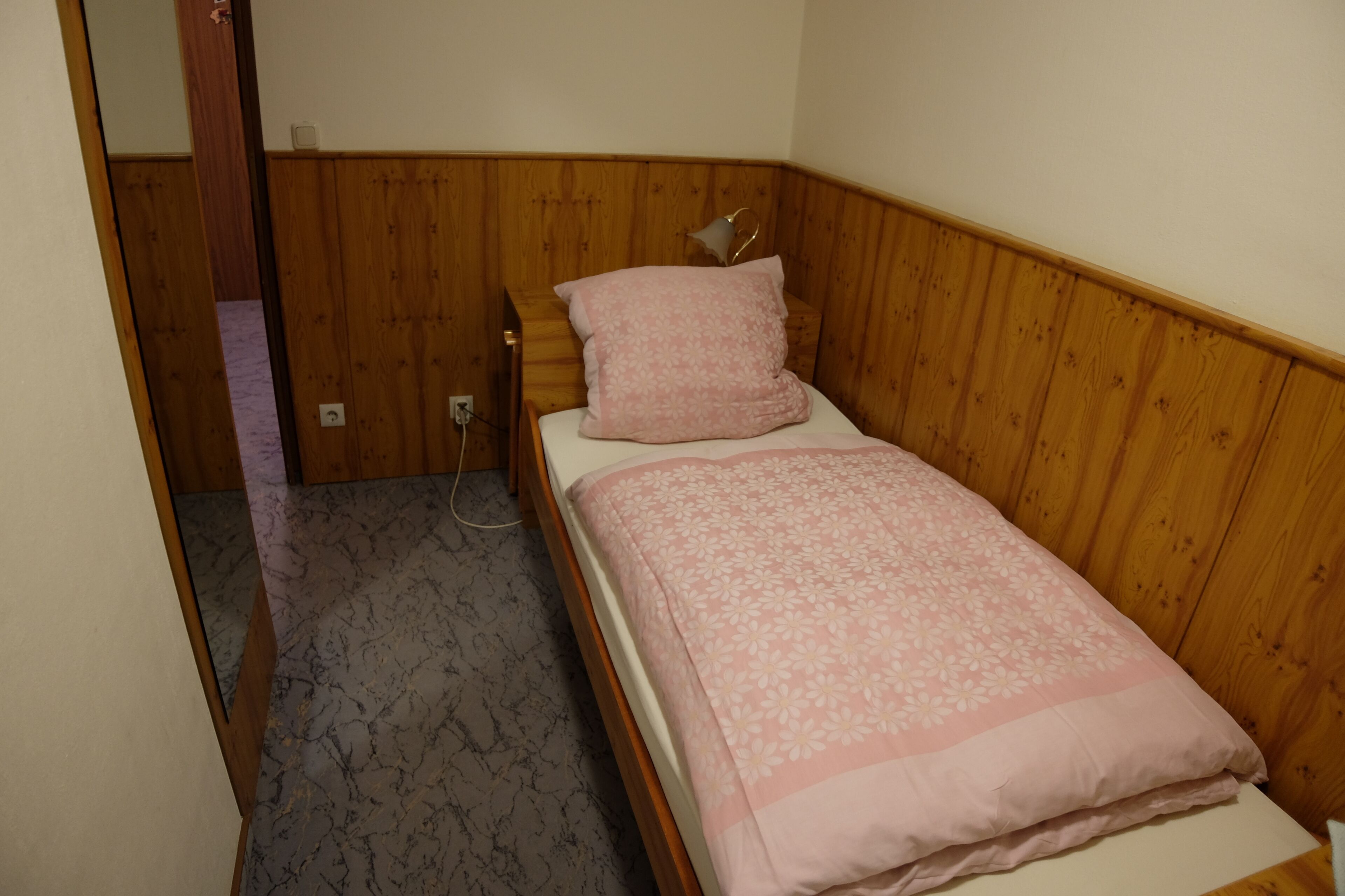 Quarto pequeno com uma cama de solteiro, painéis de madeira, colcha floral rosa e uma luminária de cabeceira.