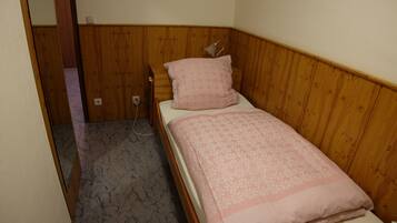 Quarto pequeno com uma cama de solteiro, painéis de madeira, colcha floral rosa e uma luminária de cabeceira.
