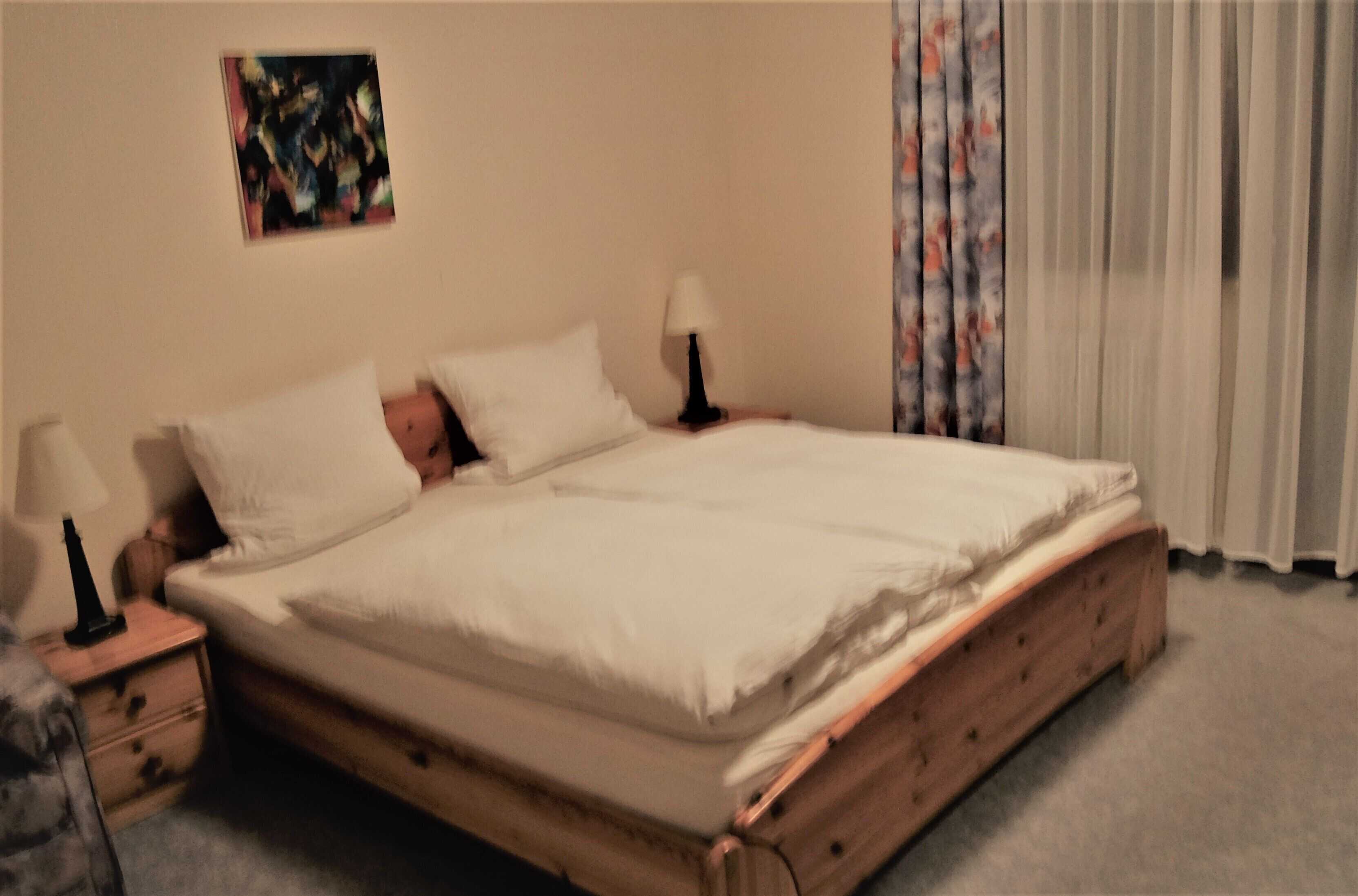 Un dormitorio con cama de madera, dos mesitas de noche, una lámpara, un cuadro en la pared y una ventana con cortinas.