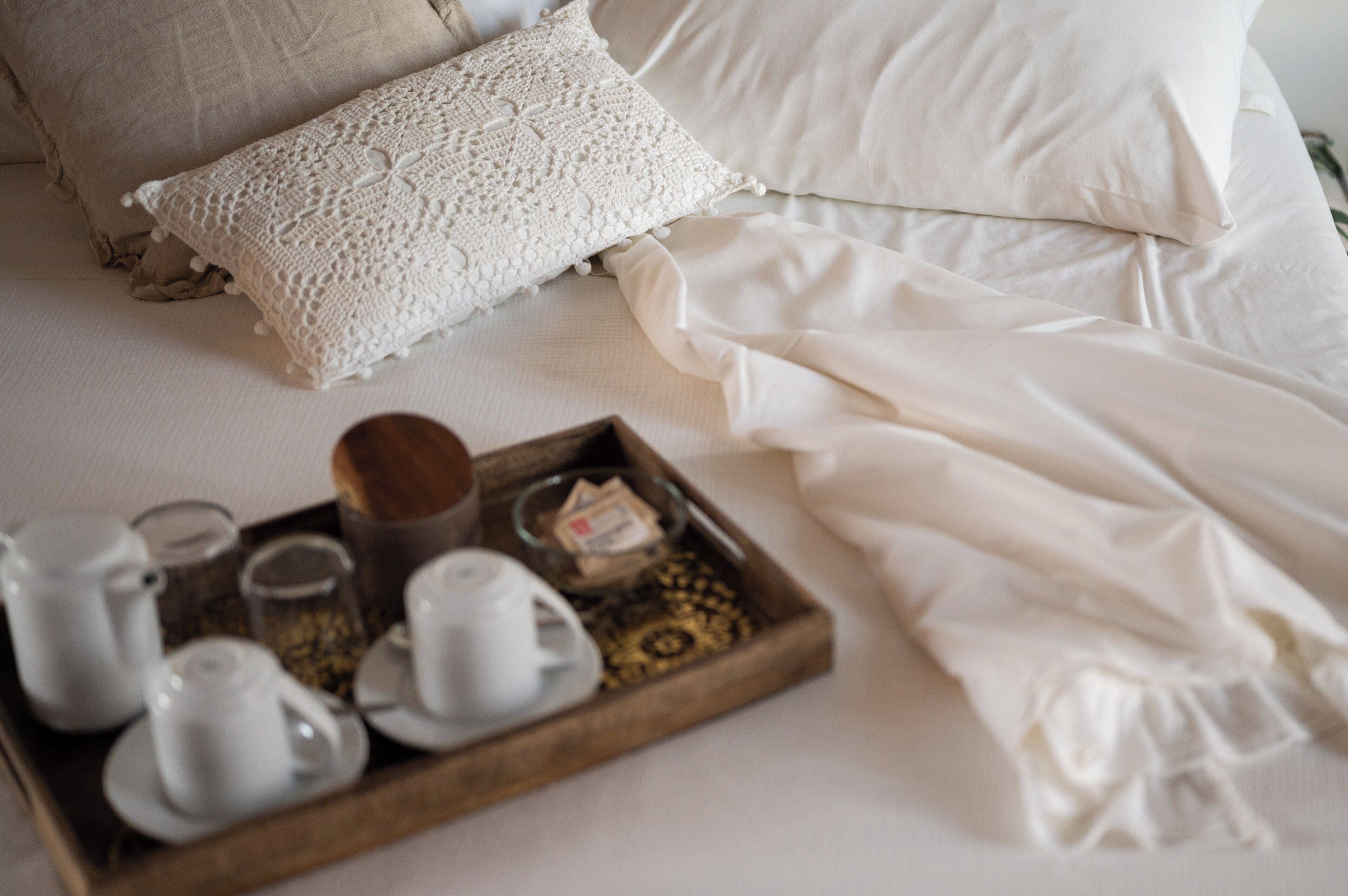 Una cama bien hecha con sábanas blancas y una almohada decorativa. Una bandeja con tazas de té y una tetera está colocada sobre la cama.