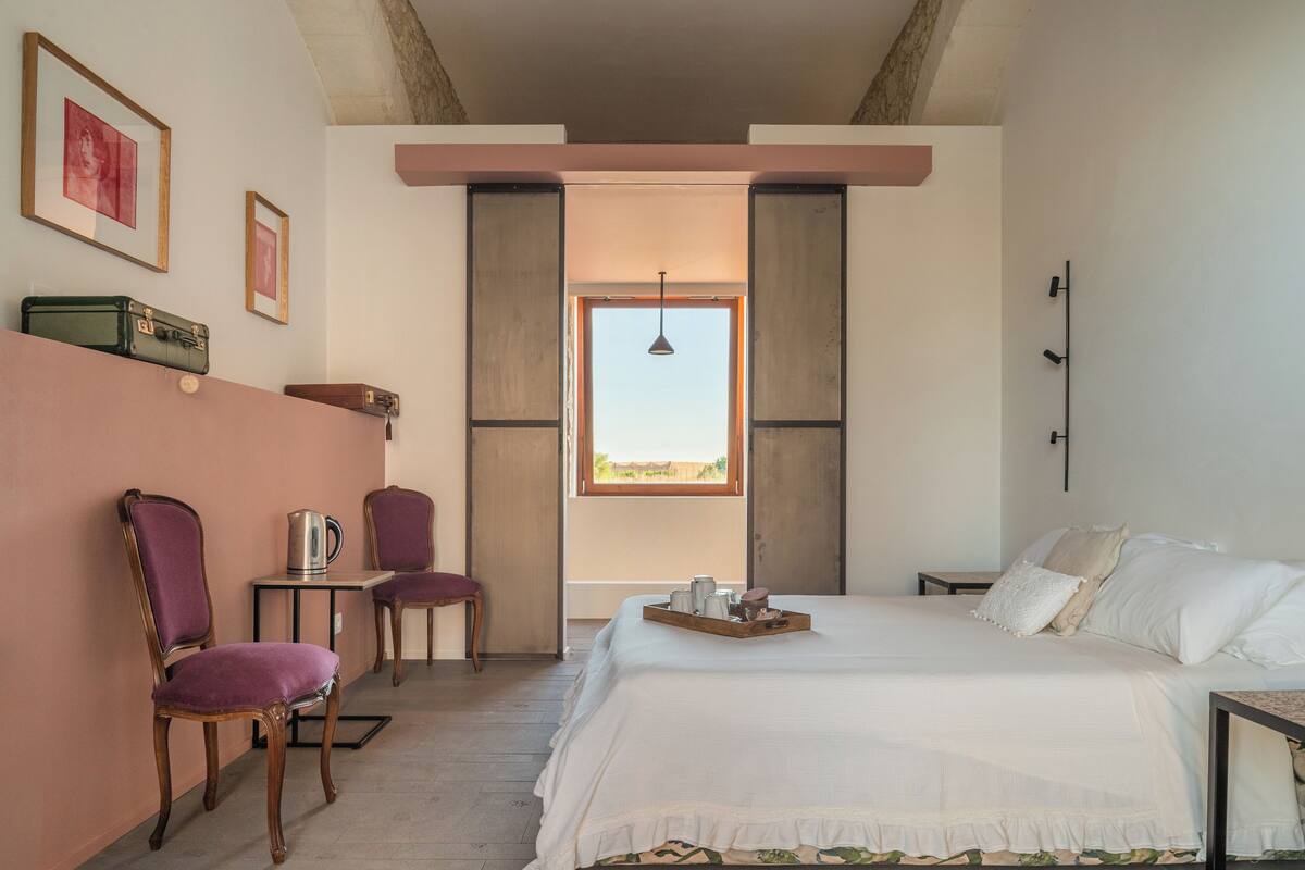 Un dormitorio con cama, dos sillones, una mesita con hervidor y una ventana con vistas.