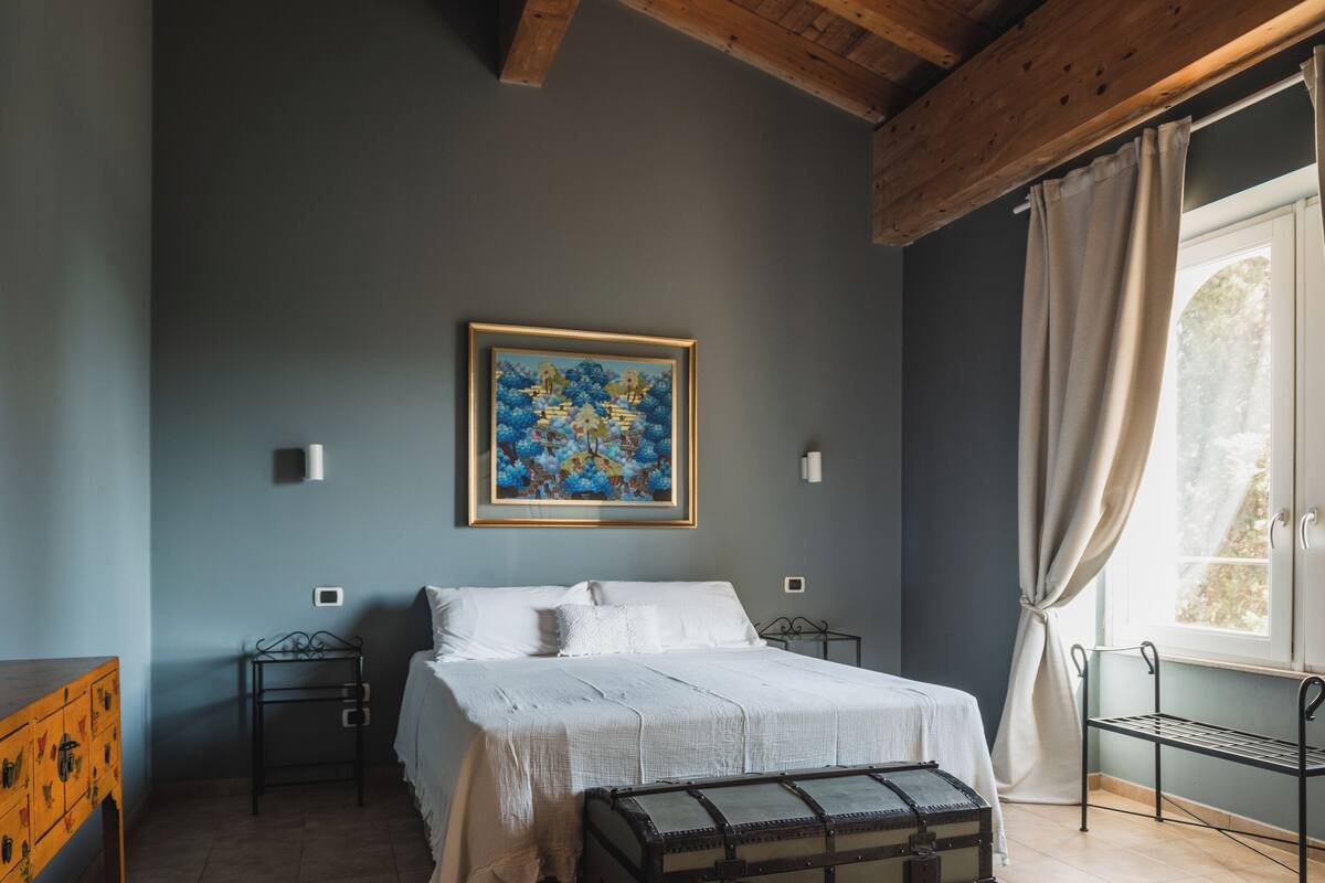Un dormitorio con cama, mesita de noche, cómoda, ventana con cortinas y un cuadro en la pared.