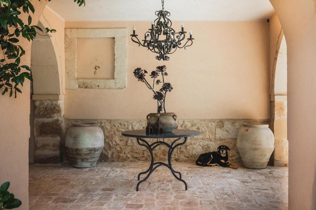 Ein rustikaler Raum mit Steinboden, einem schmiedeeisernen Tisch mit einer Vase, zwei großen Töpfen und einer hängenden, schmiedeeisernen Chandelier.