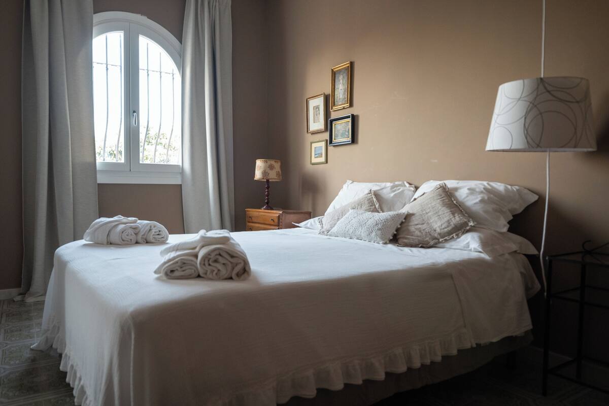Una cama bien hecha con sábanas y toallas blancas, una mesita de noche con lámpara y una ventana con cortinas.