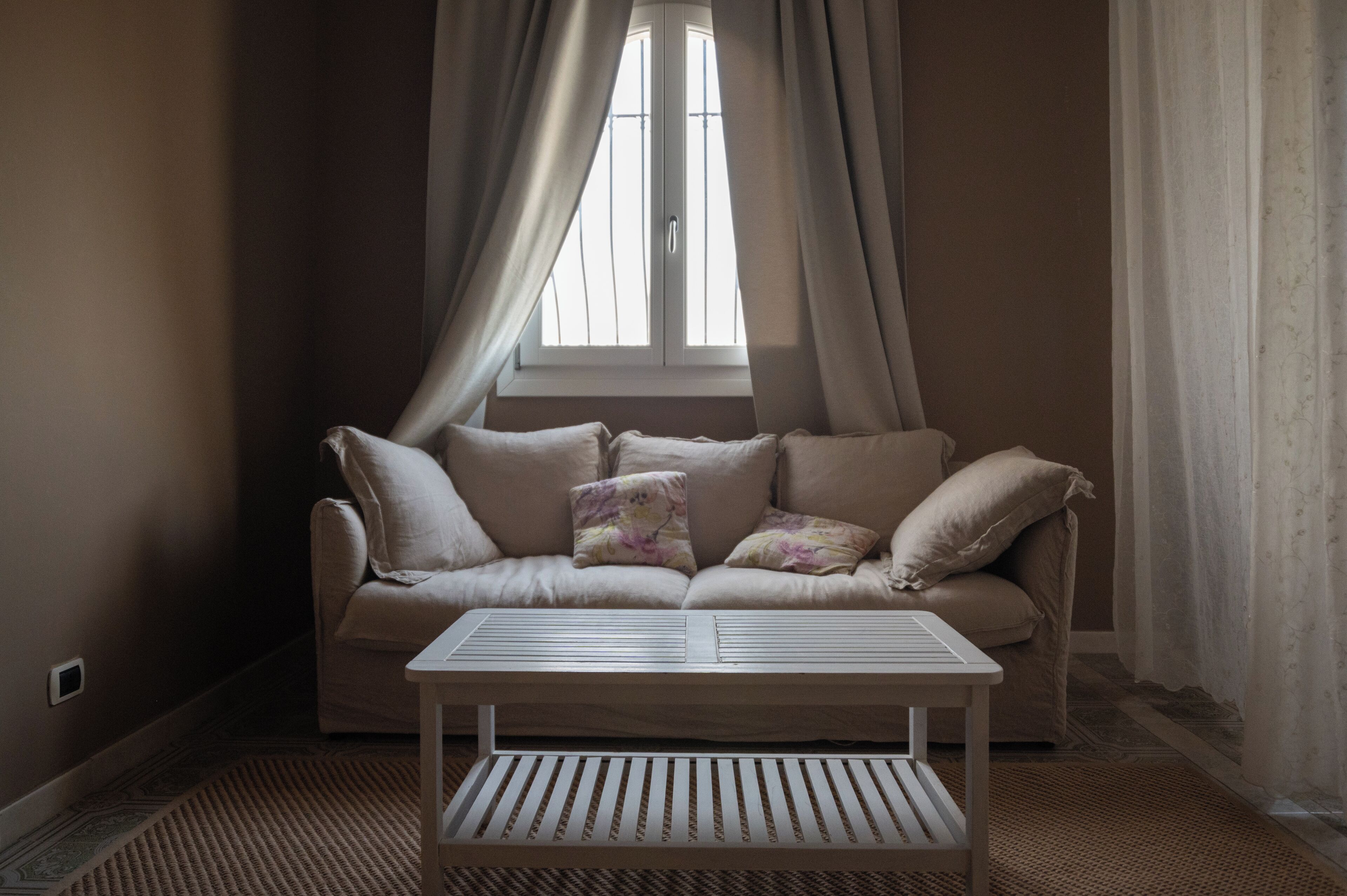 Une pièce avec un canapé beige, deux coussins, une table basse blanche et une fenêtre avec des rideaux.