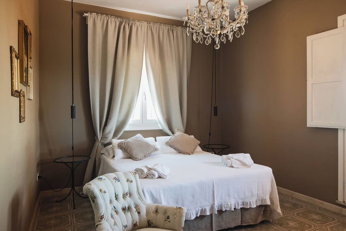 Un dormitorio con cama, cortinas, una araña de techo, una silla y un espejo fijado a la pared.