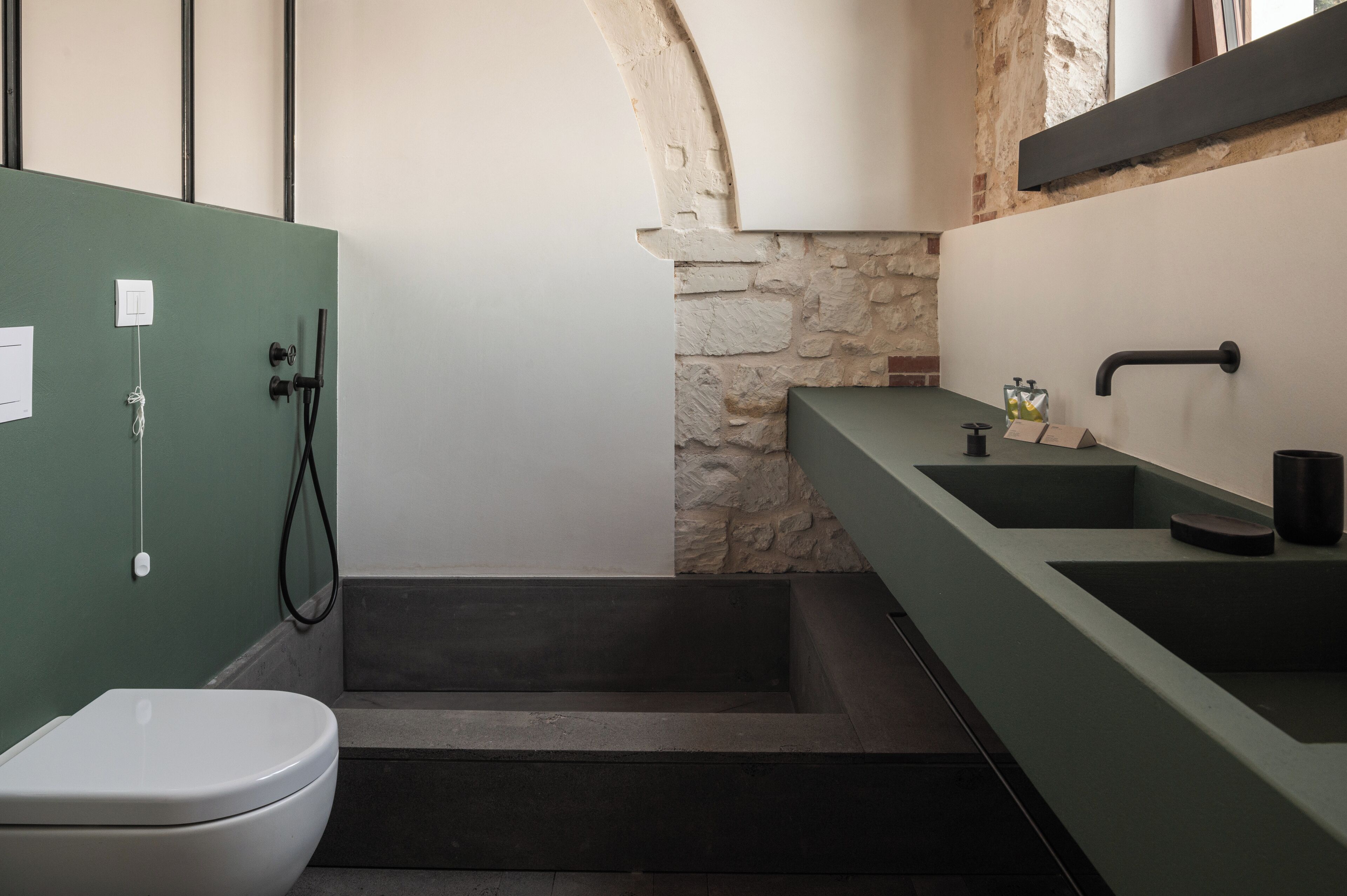A bathroom with a green wall, a white toilet, a stone accent wall, a bathtub, and a modern sink with a faucet.