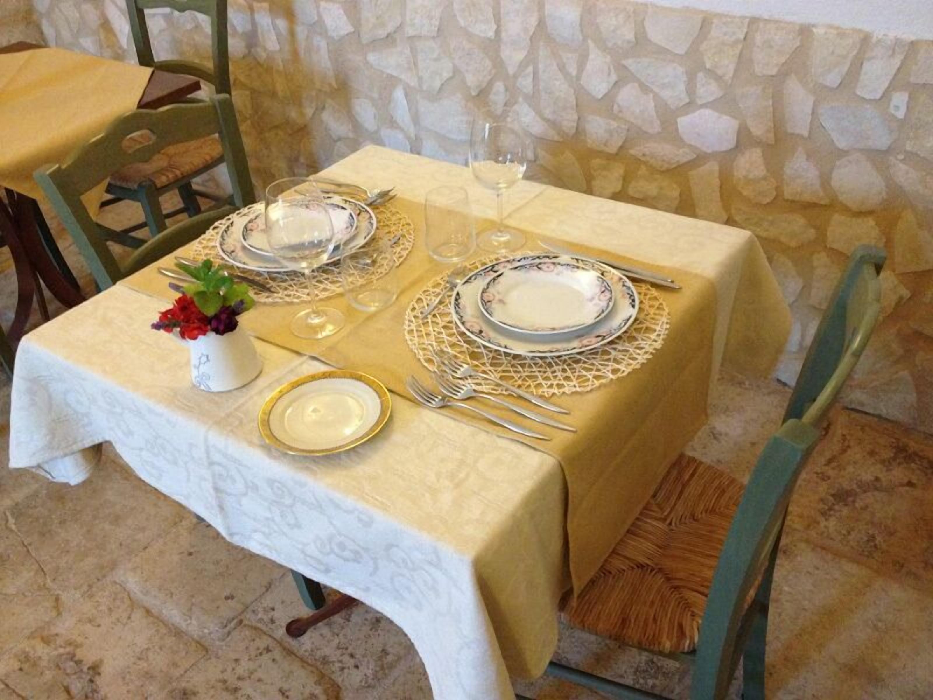 Una mesa dispuesta para una comida, con manteles blancos, platos con borde dorado y un jarrón con flores rojas.