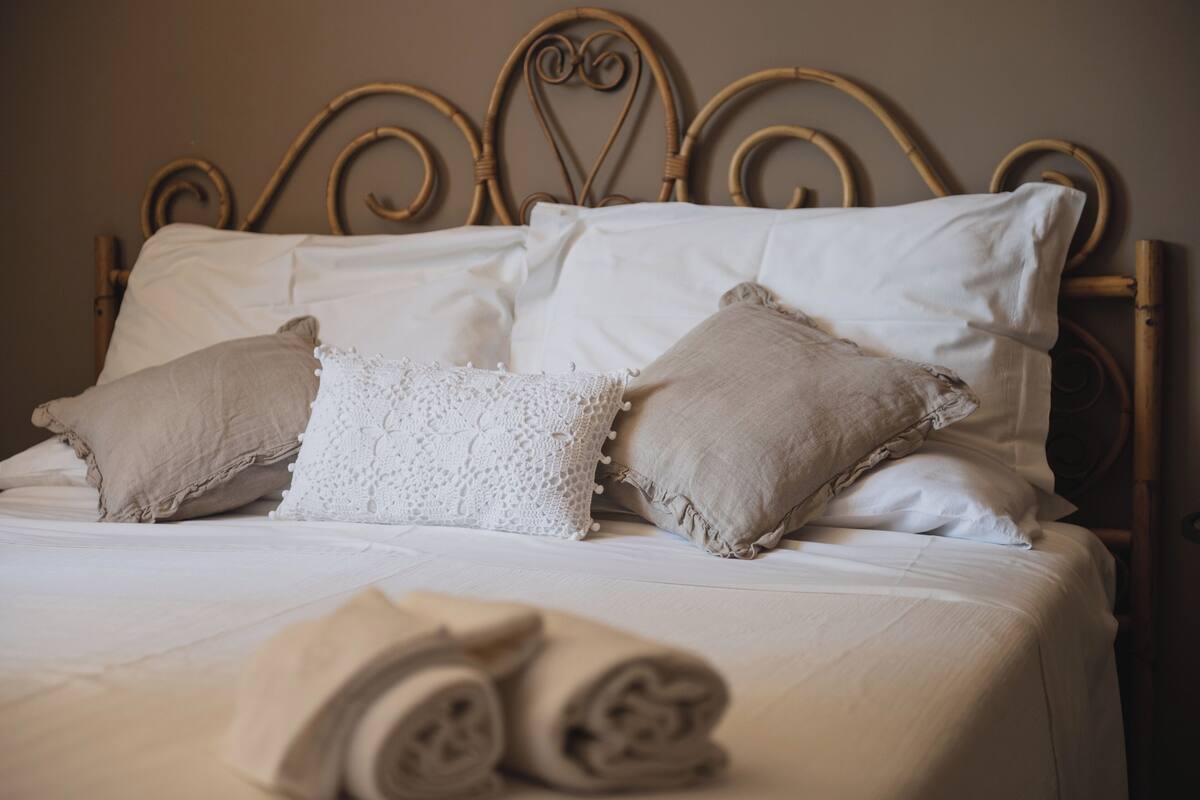 Una cama bien hecha con sábanas blancas y almohadas color beige, un cabecero de metal y toallas enrolladas sobre la cama.