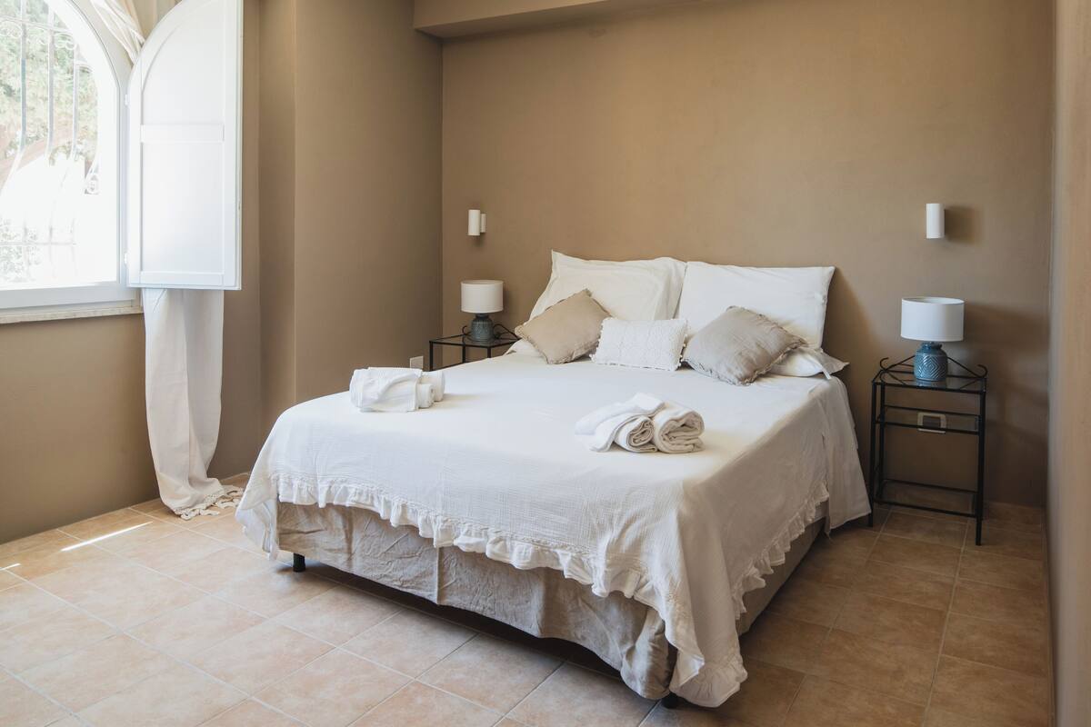 Una cama bien tendida con sábanas y almohadas blancas, dos lámparas de noche y una ventana con cortinas.