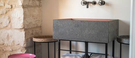 Ein Badezimmer mit einem rechteckigen Granittisch, zwei Handtuchhaltern mit gefalteten Handtüchern, einem runden Hocker und einer Steinwand.