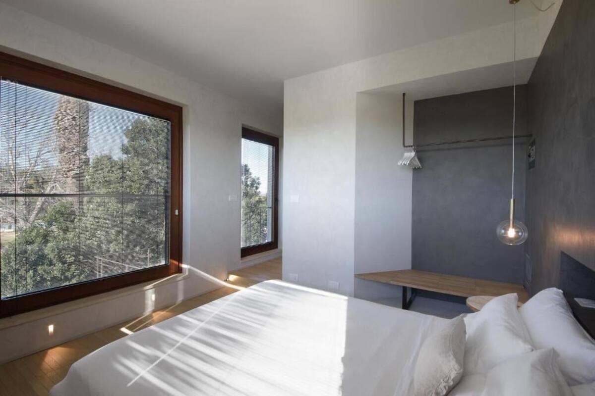 Un dormitorio moderno con un ventanal grande, una cama, un banco y una lámpara fijada a la pared.