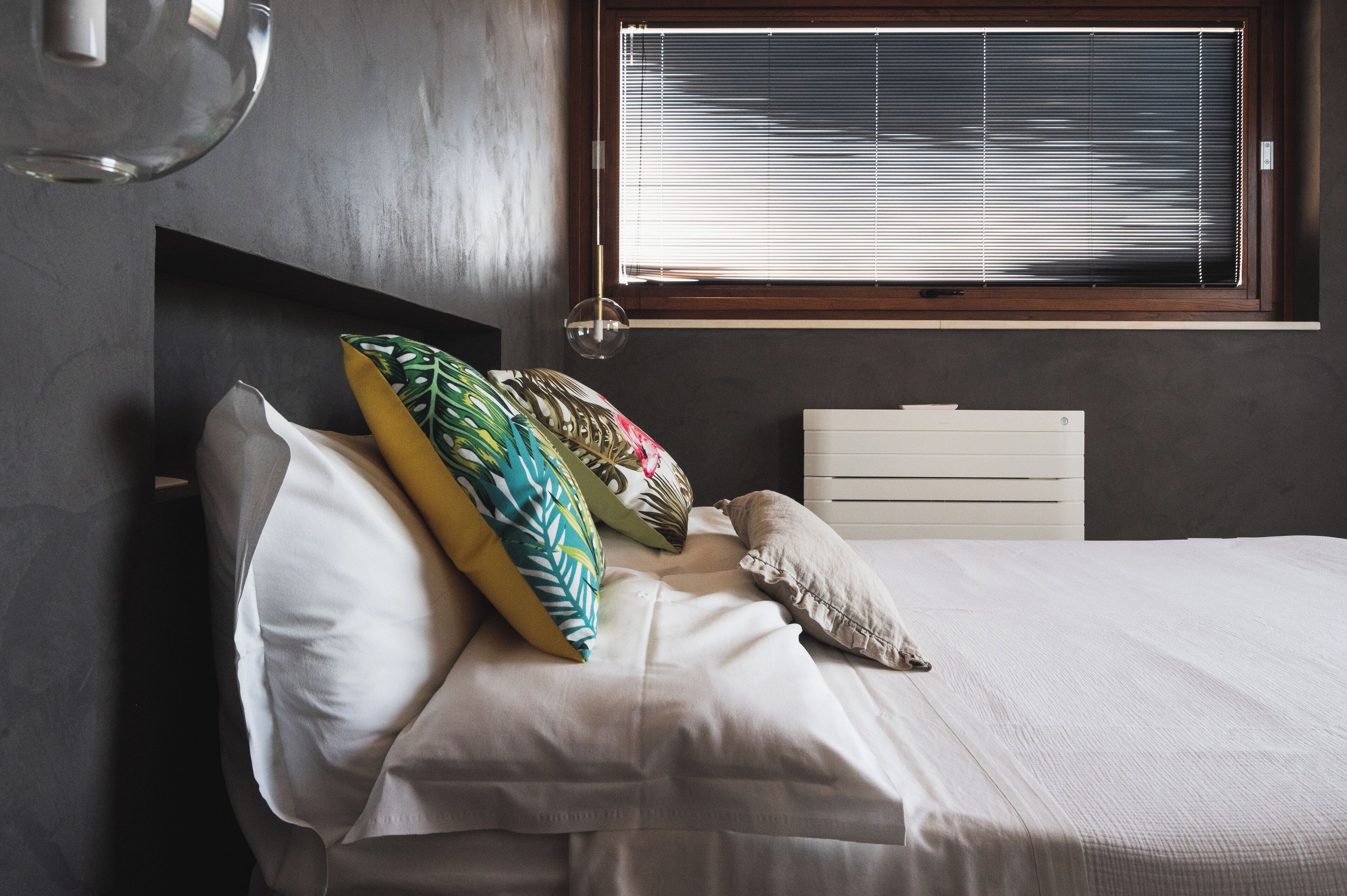 A bedroom with a bed, pillows, a bedside table, and a window with blinds.