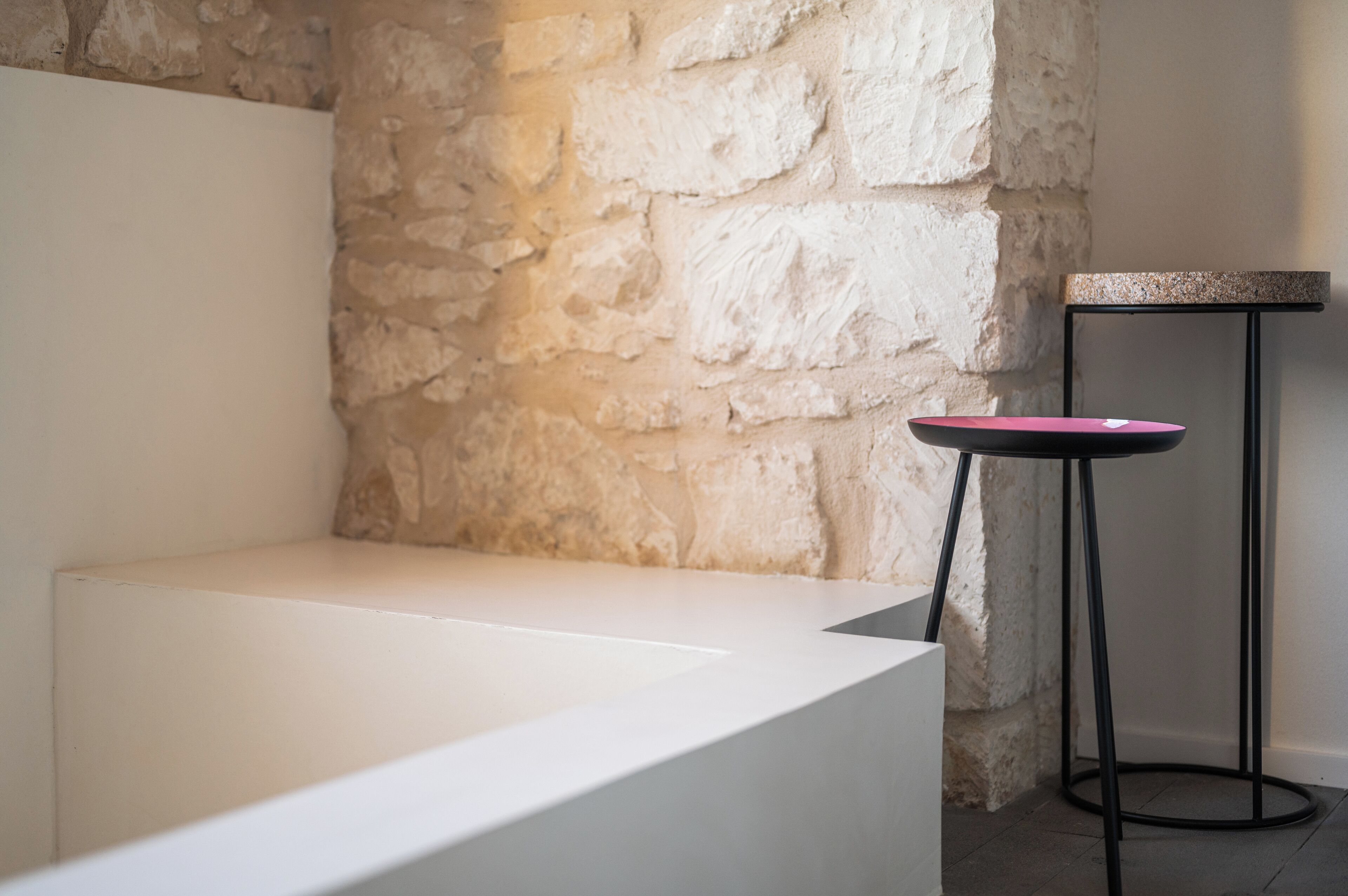 Una habitación minimalista con un banco blanco, una mesita redonda con tapa rosa y una pared de piedra.
