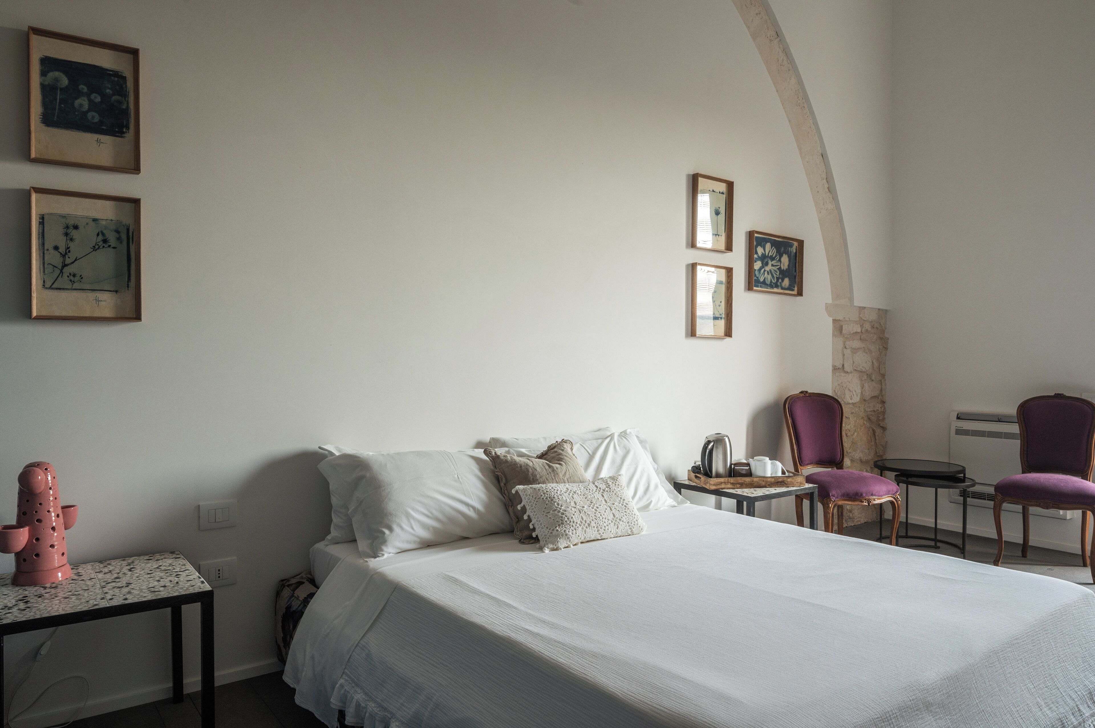 A hotel room with a bed, a small table with a kettle, two chairs, and artwork on the walls.