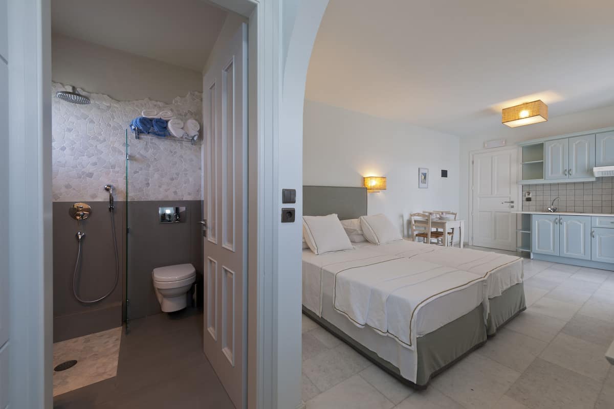 A bedroom with a bed, a bedside table, and a nightstand. There is a bathroom with a toilet, a shower, and a towel rack. The room has white walls and a tiled floor.