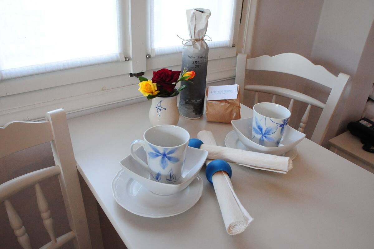 A dining table with a white tablecloth, two white cups with blue floral designs, a white vase with yellow and red flowers, a bottle labeled "Aegean Taste," and a small brown paper bag.