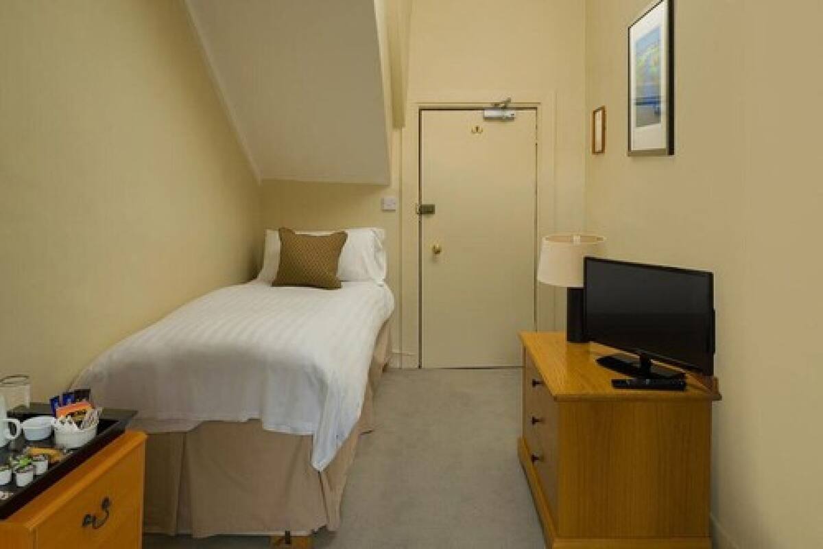 Een kleine, verzorgde slaapkamer met een enkel bed, een houten dressoir met een televisie, en een deur naar een andere kamer.