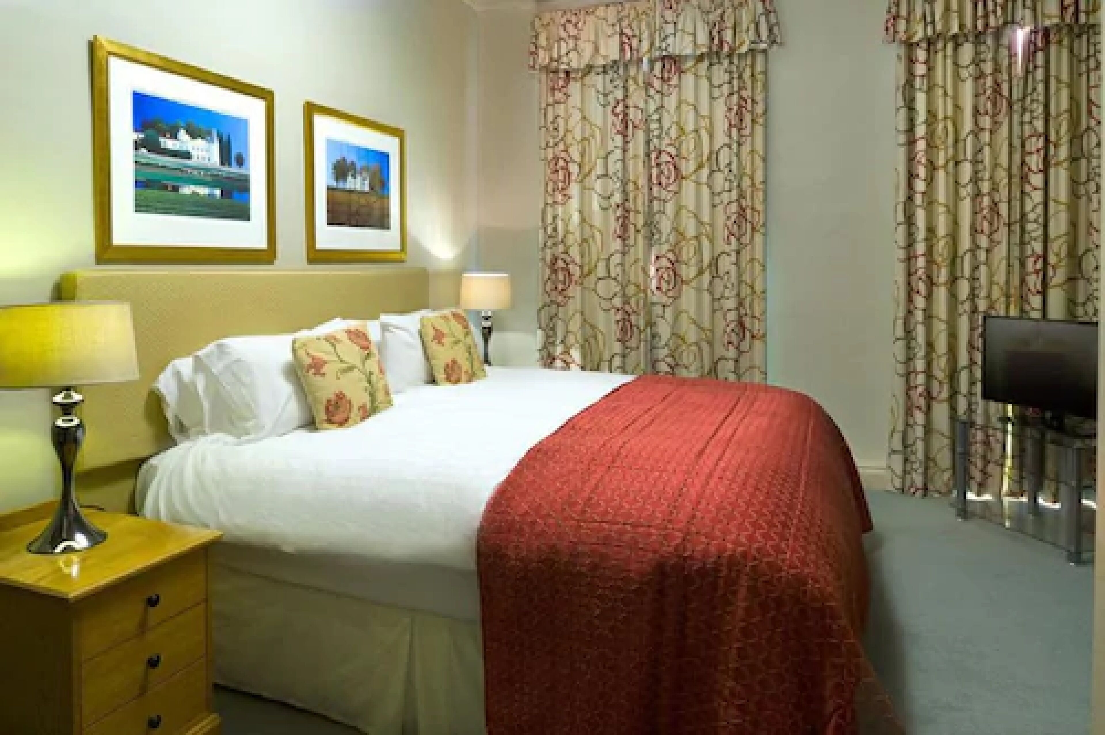 A hotel room with a bed, two bedside lamps, a TV, and two framed pictures on the wall.