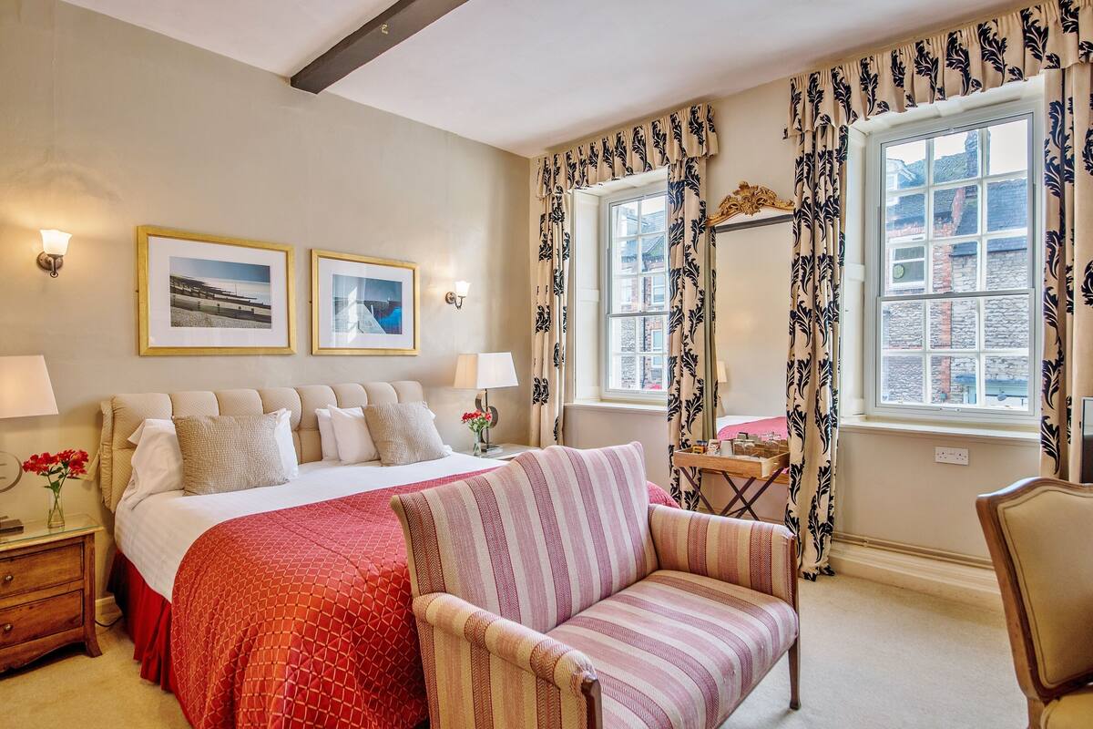 Hotelkamer met een groot bed, een gestreepte fauteuil, twee ingelijste foto’s aan de muur en een raam met gordijnen met patroon.