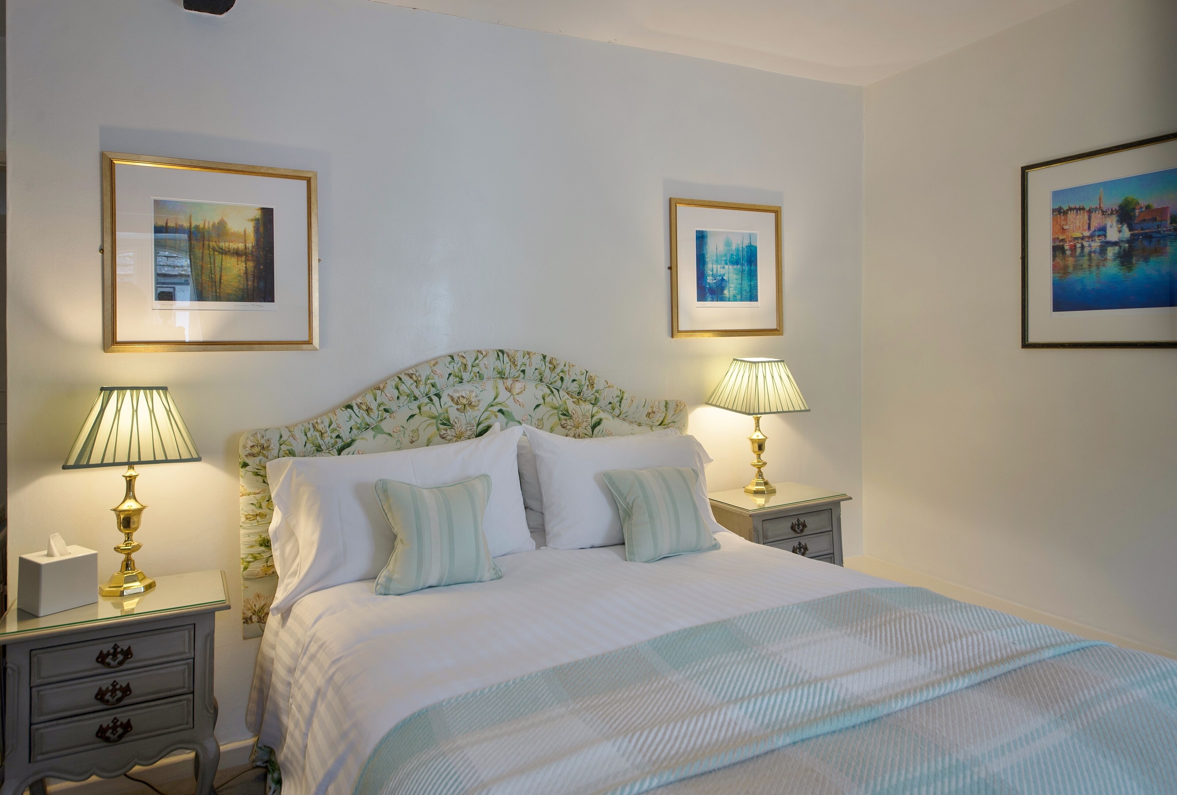A neatly made bed with a floral headboard, two bedside tables with lamps, and three framed pictures on the walls.