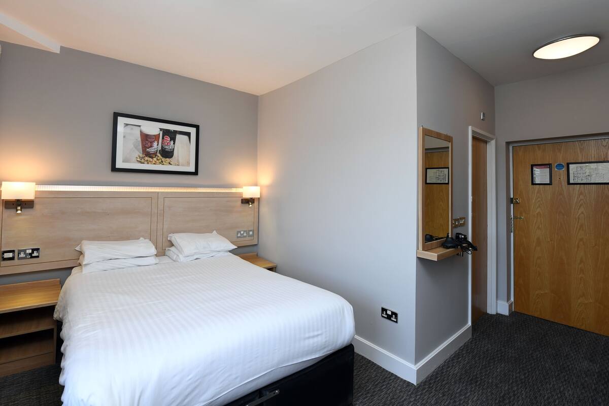 Habitación de hotel con cama, mesitas de noche, cabecero, un cuadro enmarcado, espejo, puerta y piso alfombrado.