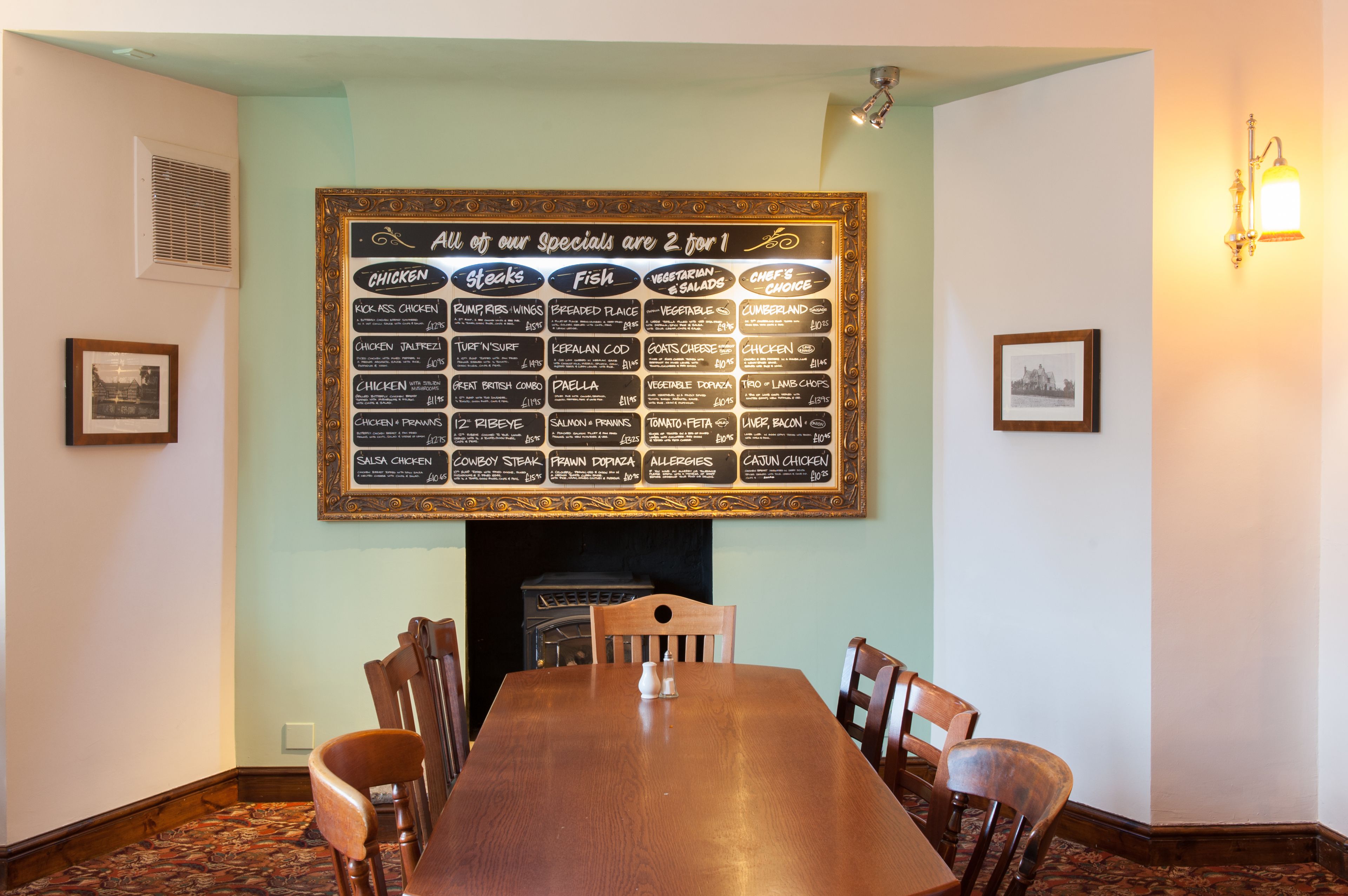 A restaurant with a large menu board above a fireplace, wooden chairs around a central table, and framed pictures on the walls.