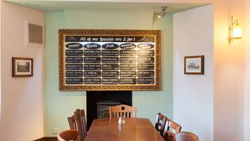A restaurant with a large menu board above a fireplace, wooden chairs around a central table, and framed pictures on the walls.