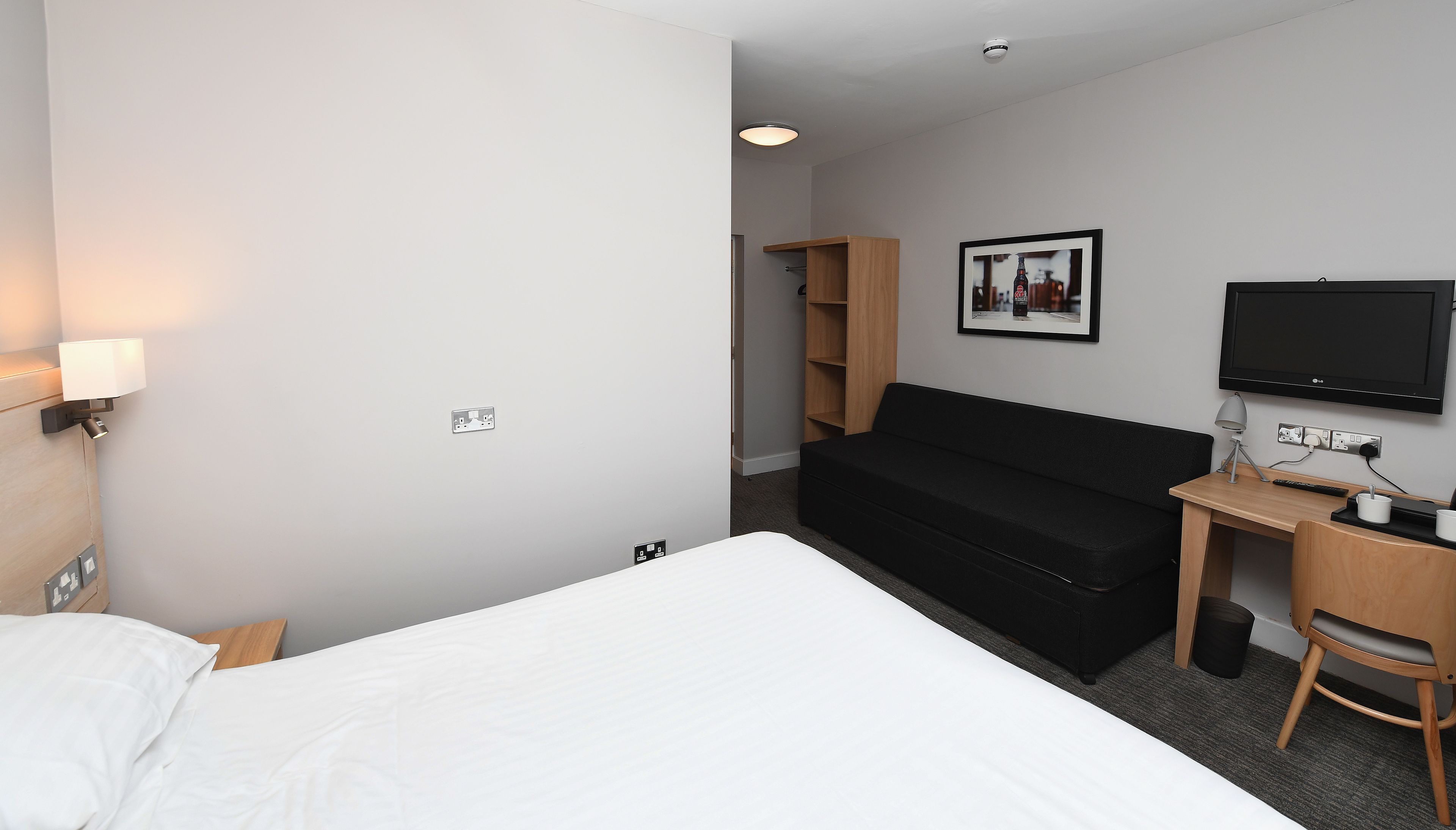 Una habitación de hotel con cama, escritorio y silla, televisor y estante con un cuadro.
