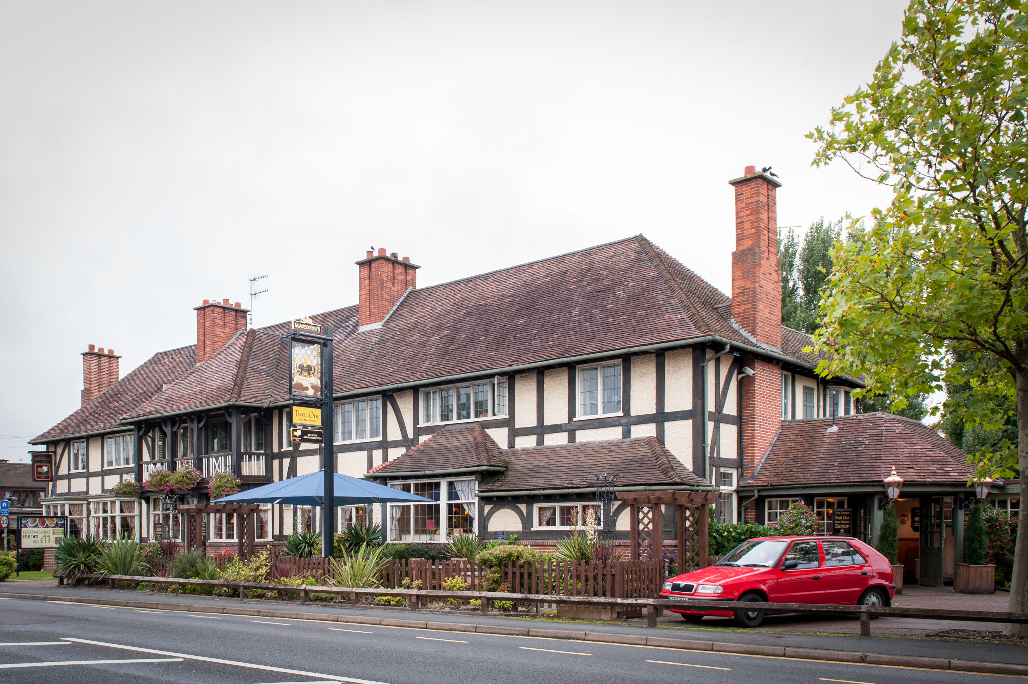A traditional pub with a Tudor-style facade, a red car parked in front, and a blue umbrella providing shade.
