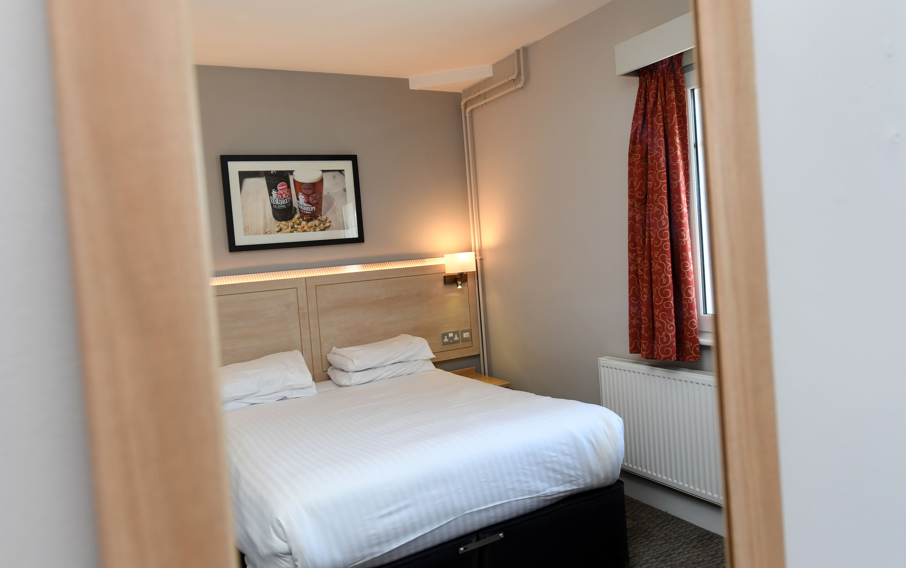 Habitación de hotel con cama, cabecera, mesita de noche, un cuadro enmarco, ventana con cortinas y un radiador.