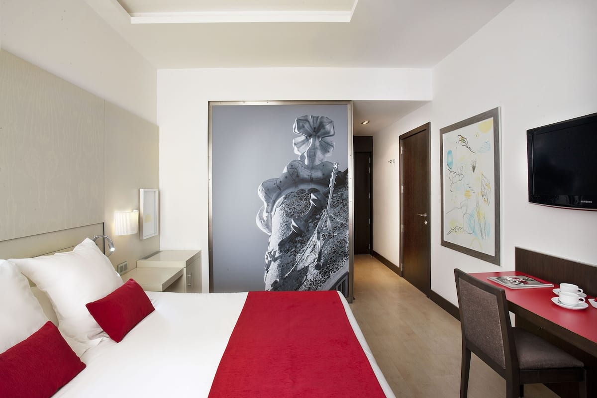 A hotel room with a bed, a desk, a chair, a TV, and artwork on the walls.