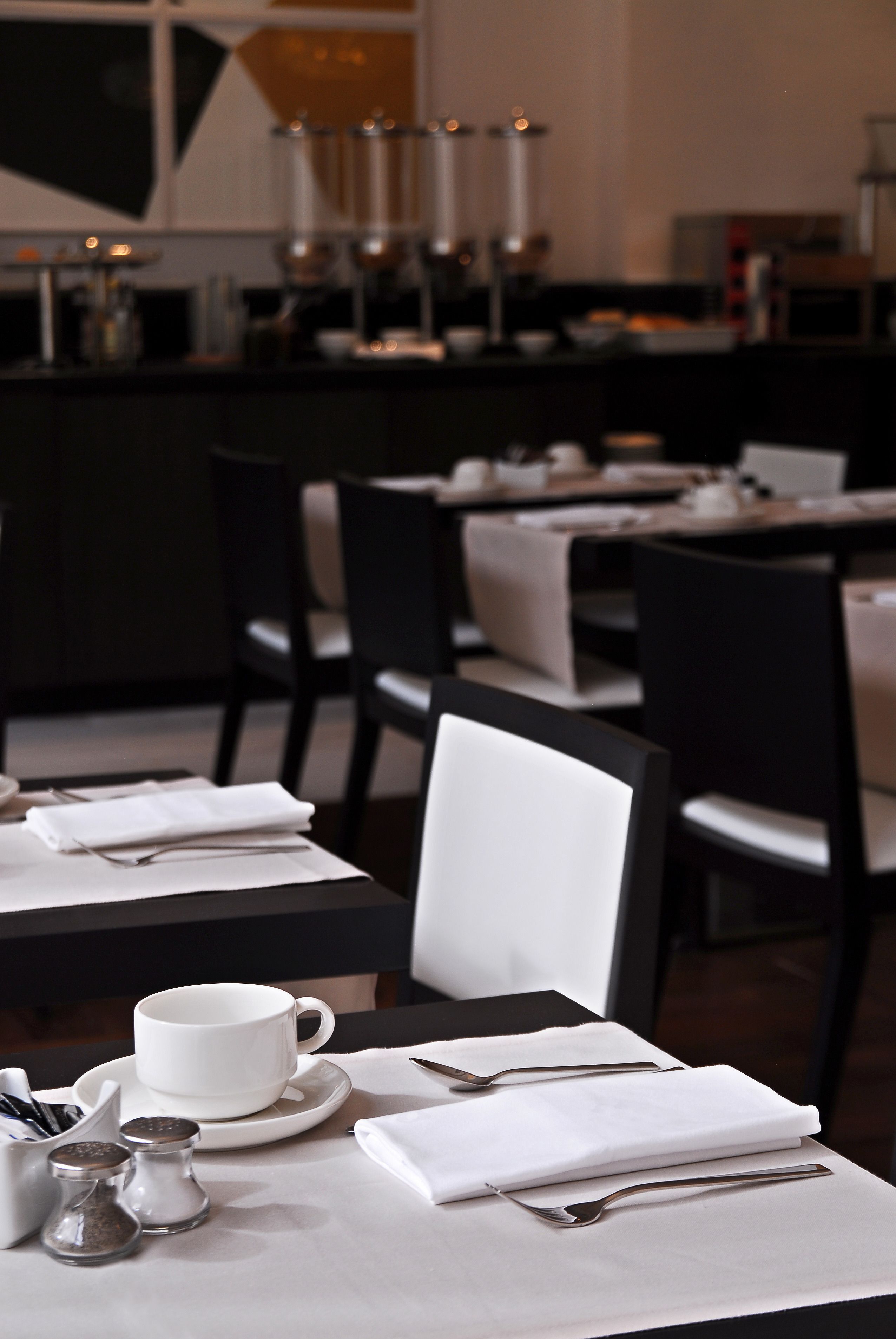 Un espace repas avec des tables dressées pour deux personnes, chacune agrémentée d’une tasse blanche, d’une soucoupe et de couverts.