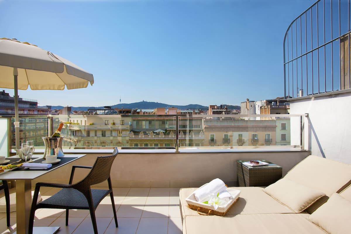 A balcony with a table, chairs, and a sofa. There is a view of buildings and a mountain in the background.