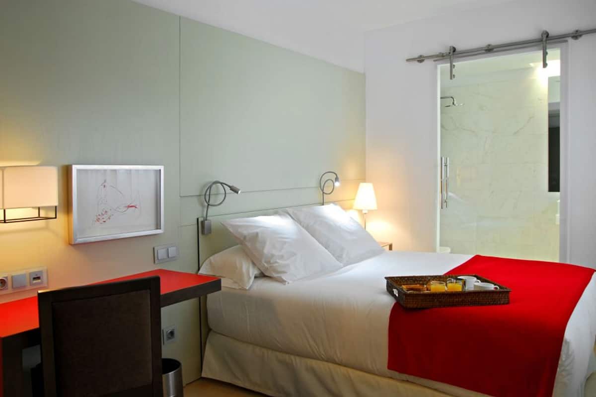 A hotel room with a bed, a desk, a chair, a lamp, a framed picture, and a bathroom with a shower.