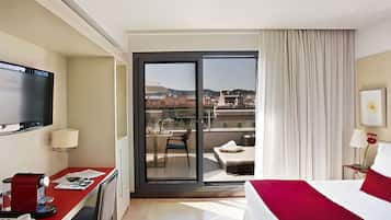 A hotel room with a bed, a desk with a coffee maker, a TV, a balcony with a chair, and a view of buildings and mountains.