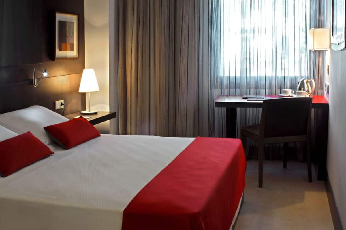 A hotel room with a bed, a desk, a chair, a lamp, and a window with curtains.