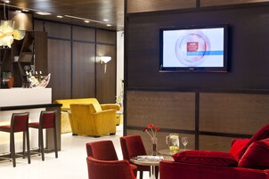A modern hotel lobby with a bar area, red and yellow seating, and a television displaying the BBC World News Business channel.