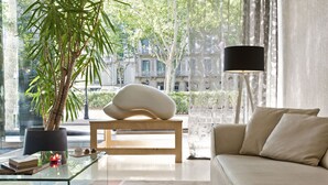 Una sala de estar moderna con una mesa de centro de vidrio, un sofá color beige, una planta grande en maceta y vistas de un paisaje urbano a través de grandes ventanales.