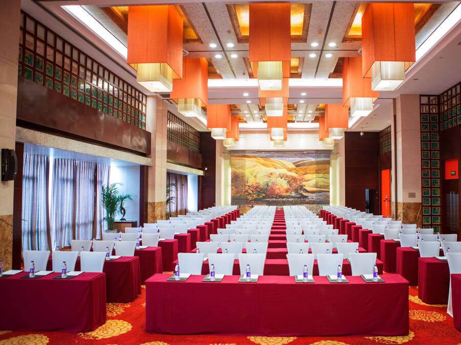 A conference room with rows of white chairs, red tables, and a large screen at the front.