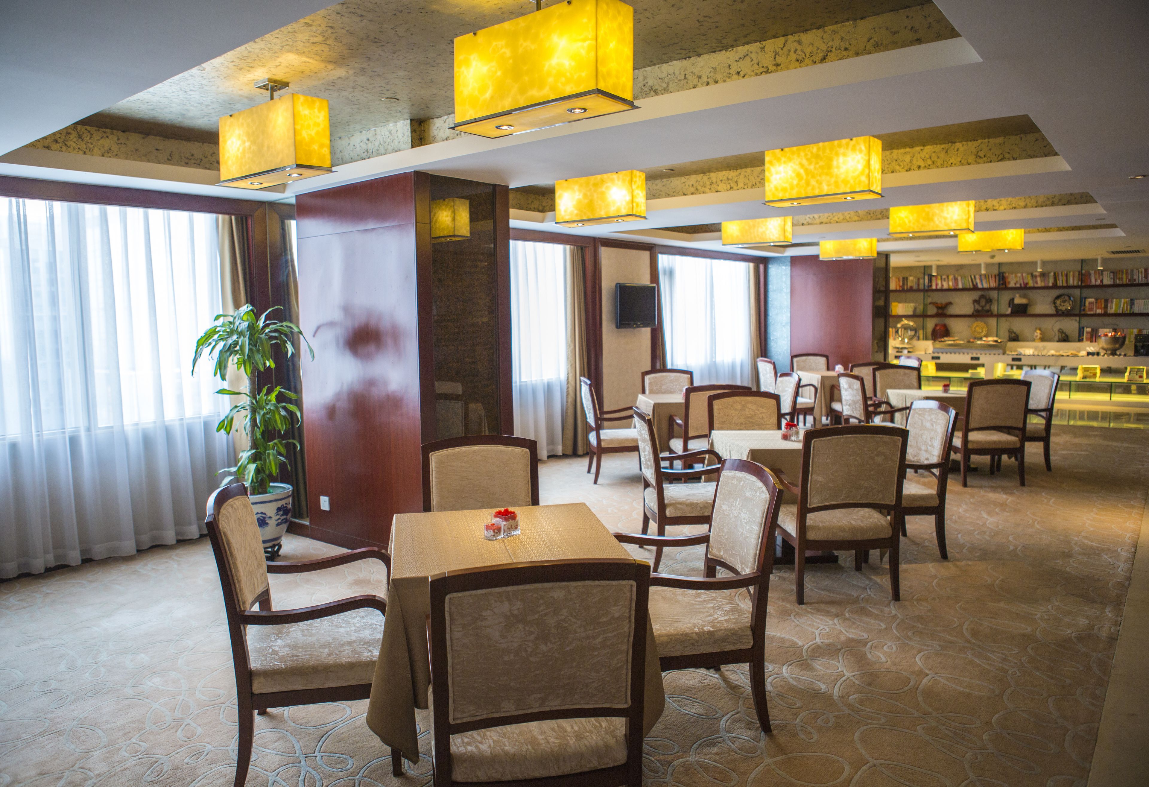 A hotel lobby with multiple dining tables and chairs, a large potted plant, and a bookshelf filled with books.