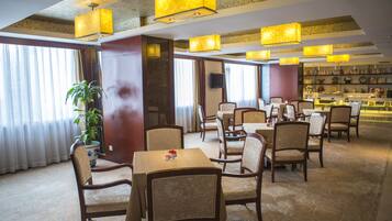 A hotel lobby with multiple dining tables and chairs, a large potted plant, and a bookshelf filled with books.