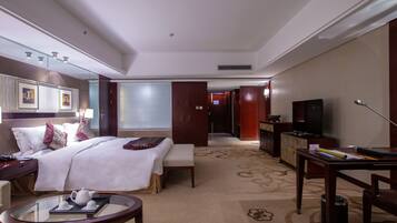 A hotel room with a bed, bedside table, lamp, TV, and a desk with a chair.
