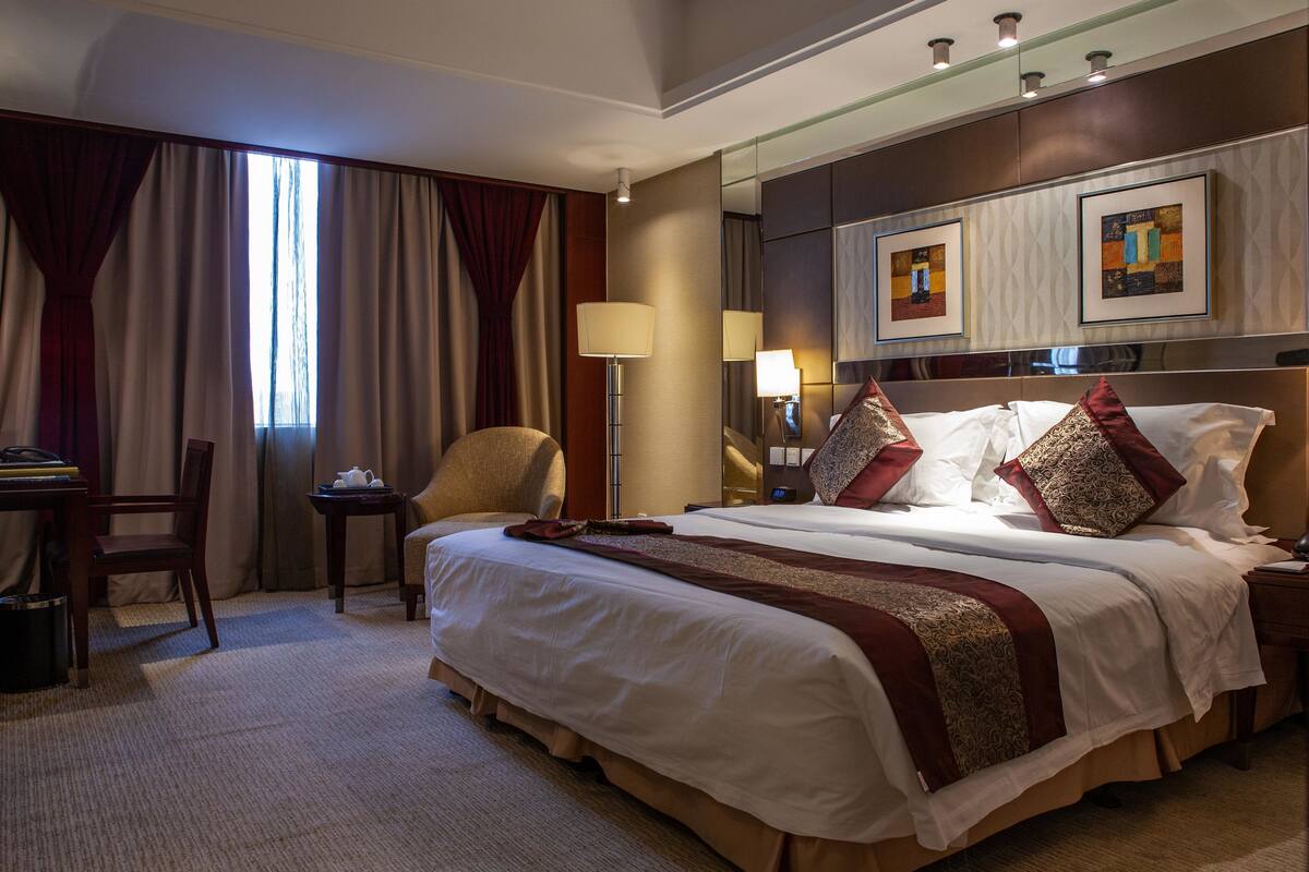A hotel room with a large bed, a desk, a chair, a lamp, and artwork on the wall.