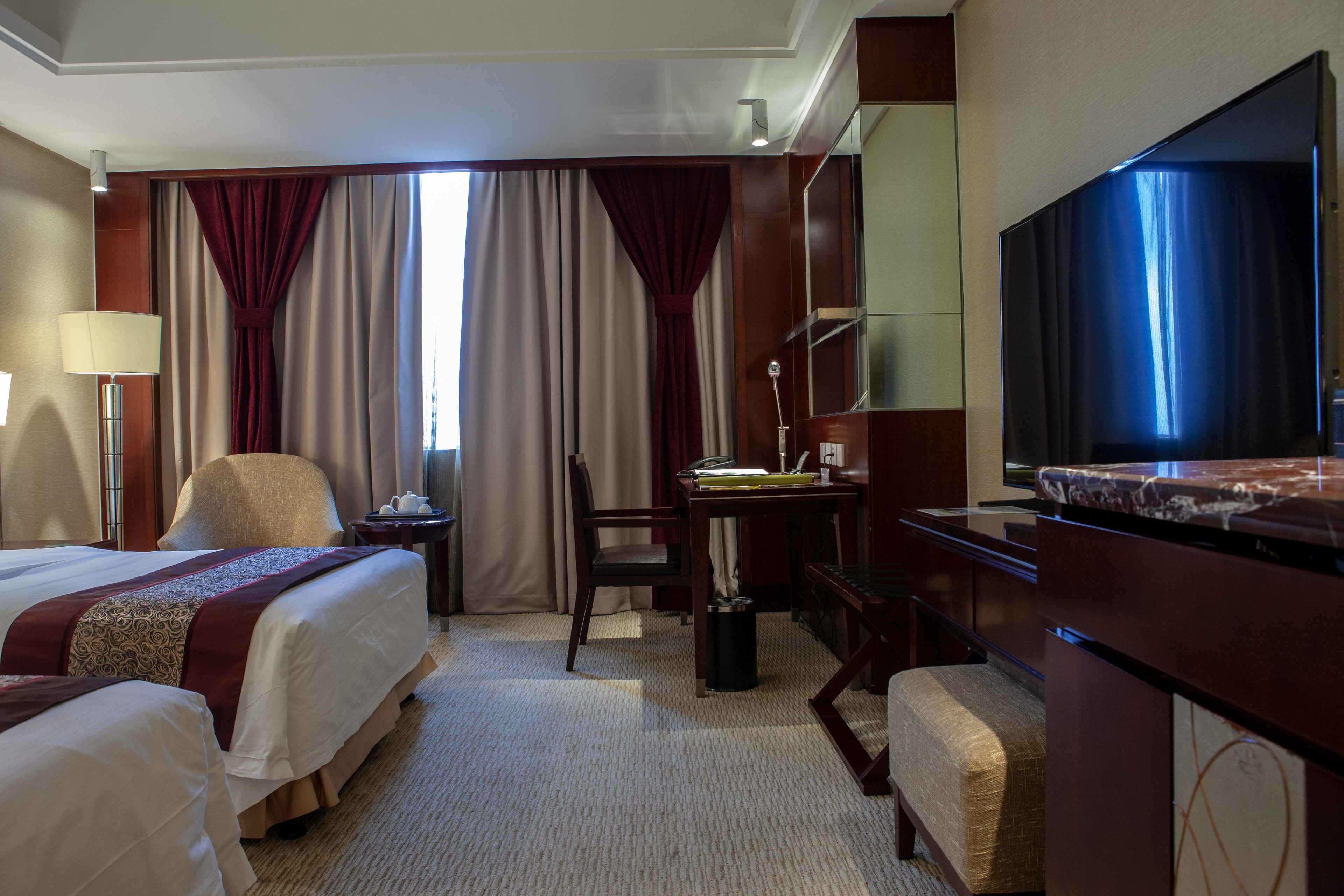 A hotel room with two beds, a desk, a chair, a TV, and a large window with curtains.