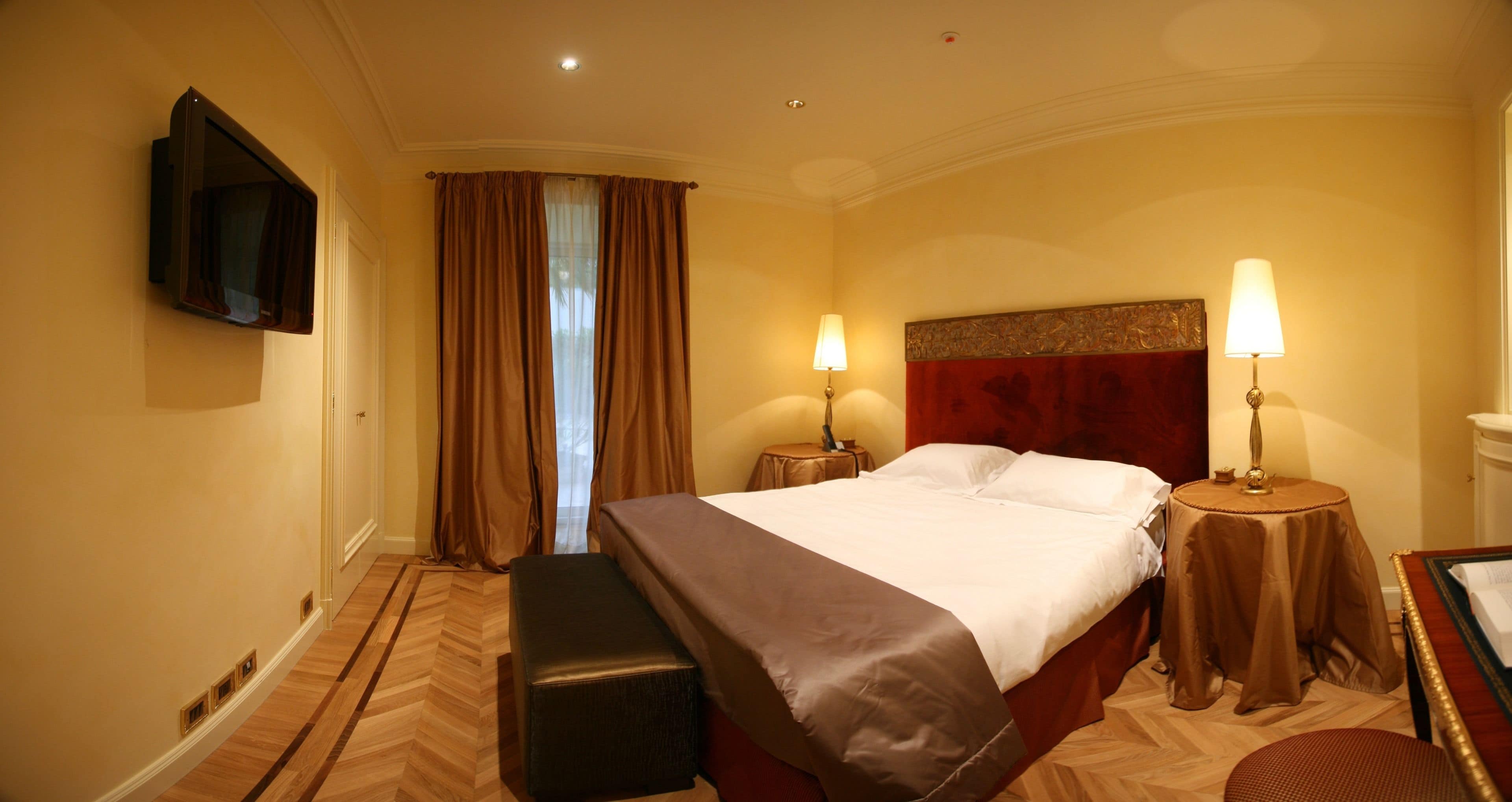 A hotel room with a bed, two bedside tables with lamps, a bench, a flat-screen TV mounted on the wall, and a window with curtains.