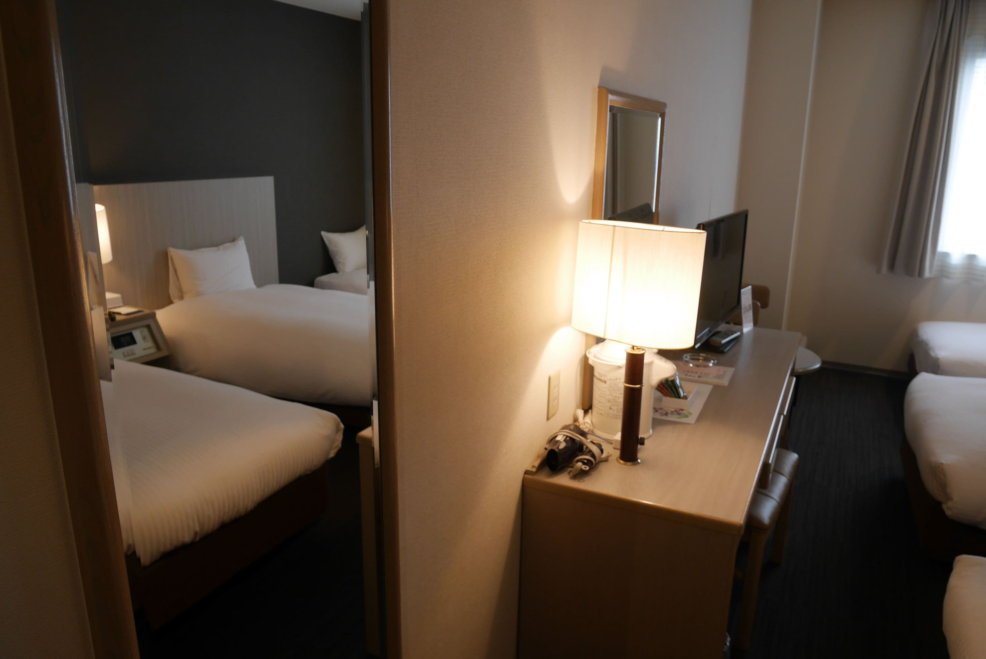 A hotel room with two beds, a desk with a lamp, a mirror, and a window with curtains.