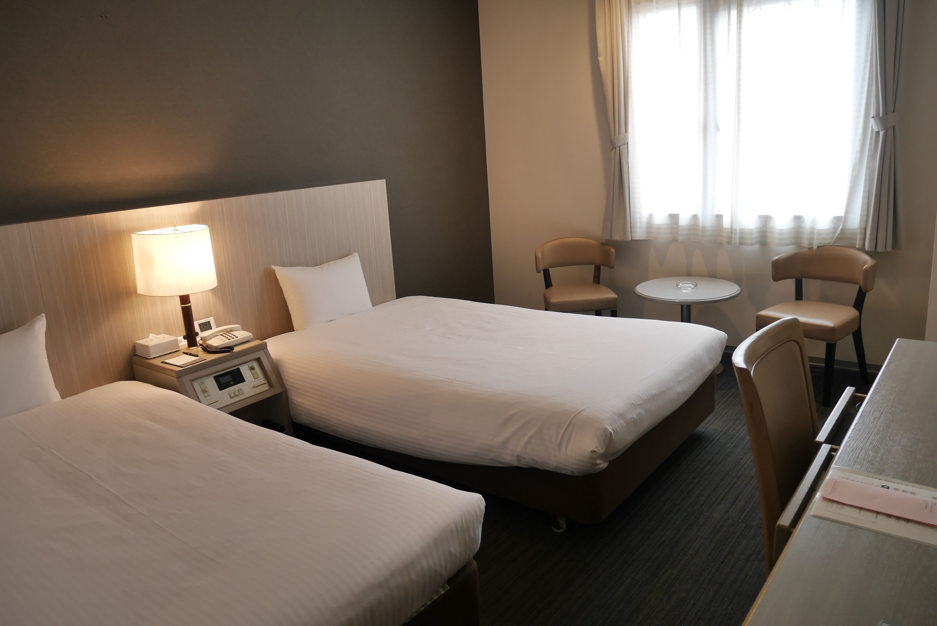 A hotel room with two beds, a bedside table with a lamp, a small round table, two chairs, and a desk with a chair.