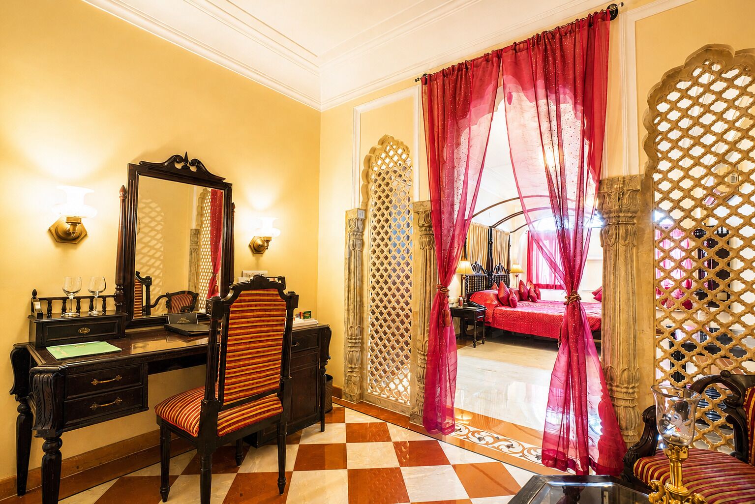 A room with a checkered floor, a desk with a mirror, a chair, and a bed with red curtains.